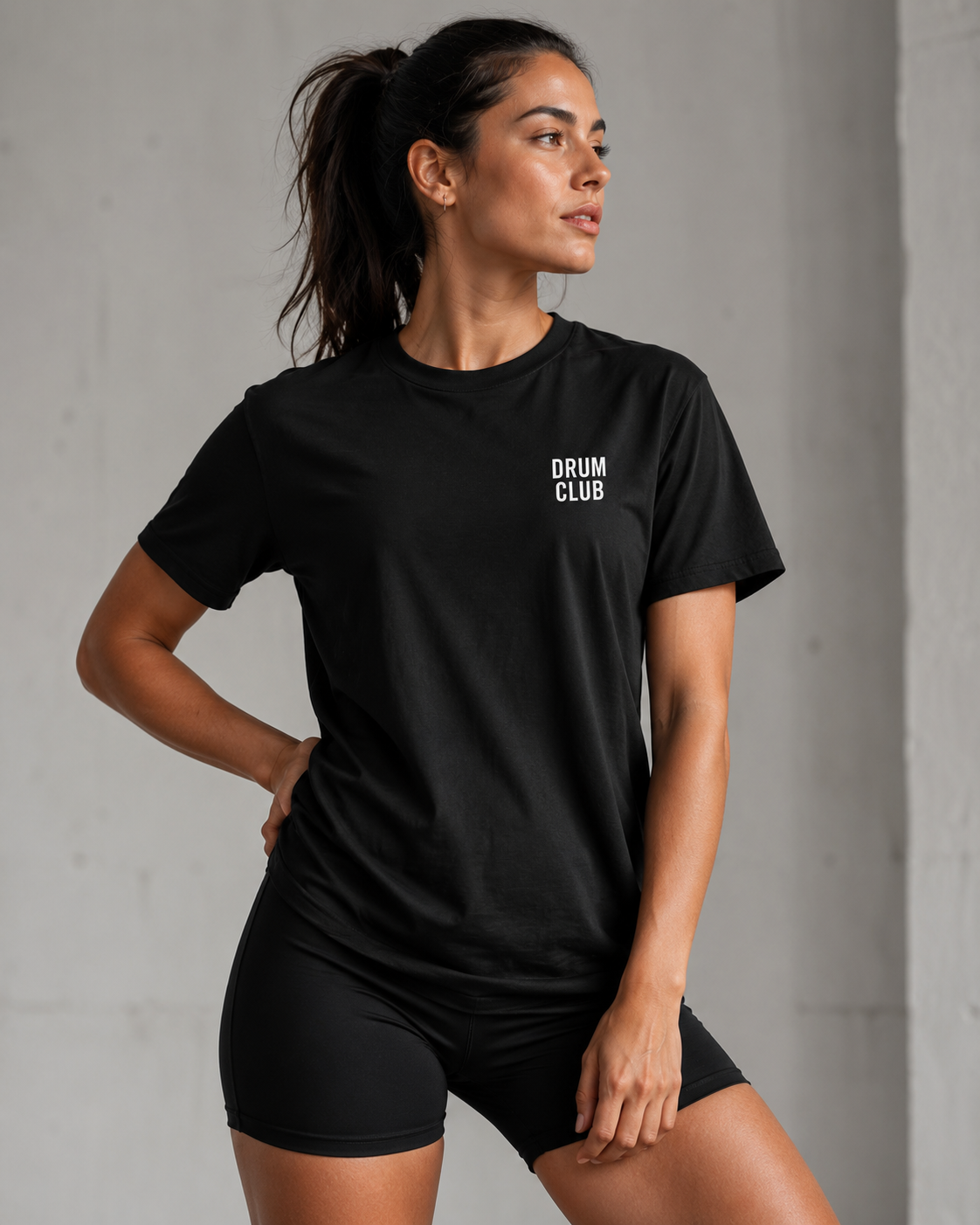 Athletic woman wearing a black DRUM CLUB T-shirt with a white left chest logo, styled with black shorts against a clean neutral studio background.